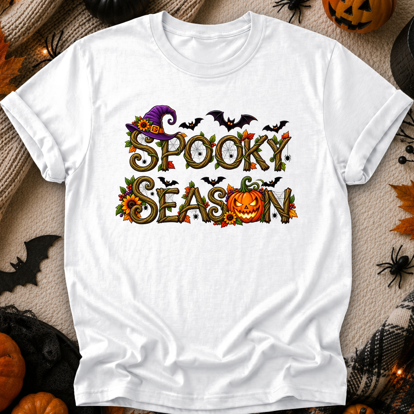 A white short-sleeve graphic T-shirt laid flat on a cozy beige fabric background with Halloween props. The shirt features a colorful 'Spooky Season' design with a purple witch hat, bats, clocks, leaves, sunflowers, and a jack-o'-lantern.