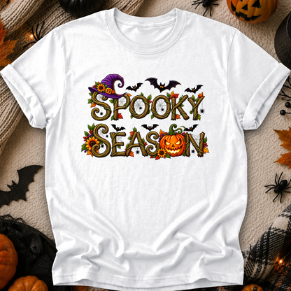 A white short-sleeve graphic T-shirt laid flat on a cozy beige fabric background with Halloween props. The shirt features a colorful 'Spooky Season' design with a purple witch hat, bats, clocks, leaves, sunflowers, and a jack-o'-lantern.