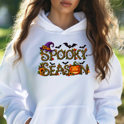 A woman wearing a white pullover hoodie with a colorful 'Spooky Season' graphic on the chest, featuring a witch hat, bats, sunflowers, and a pumpkin.