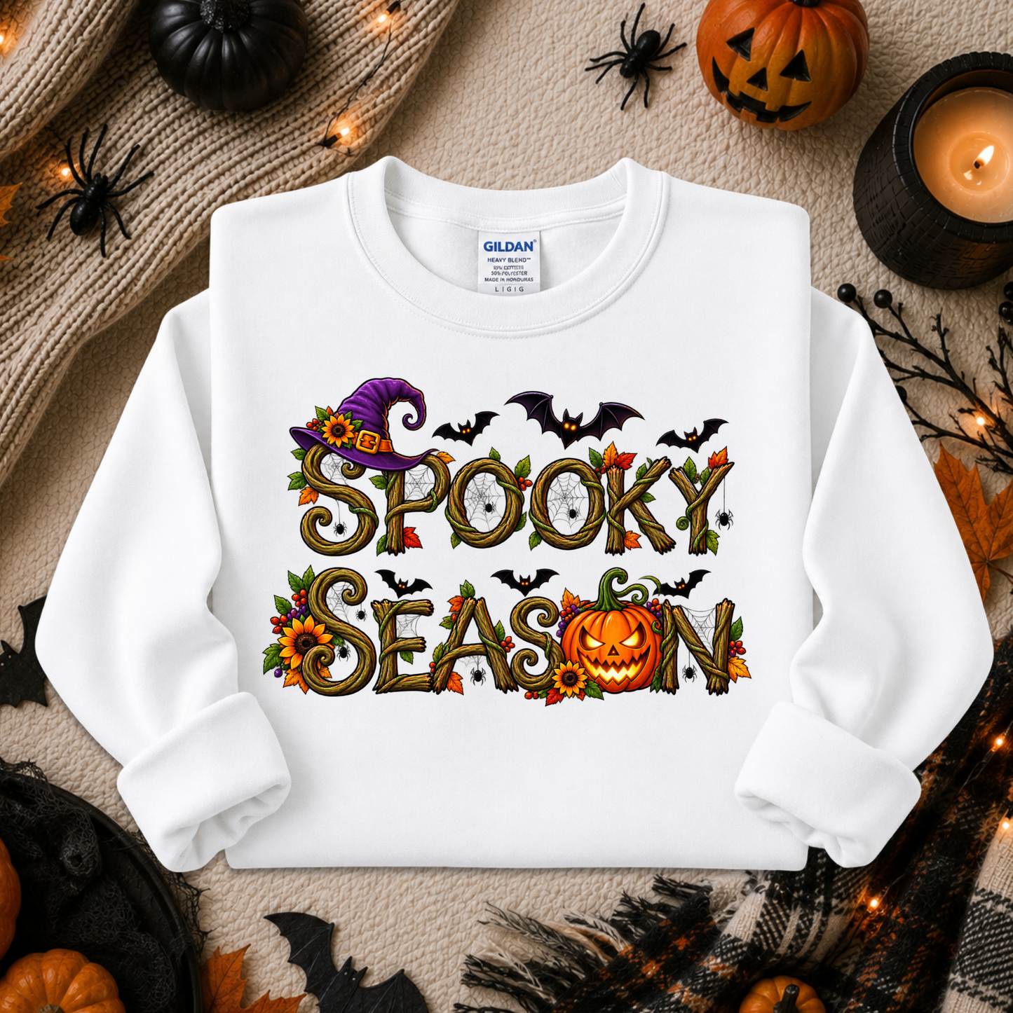 A white long-sleeve crewneck sweatshirt with a 'Spooky Season' graphic, laid flat on a neutral background with Halloween props including pumpkins, a candle, and spider decorations. The brand Gildan is visible on the inner neck label.
