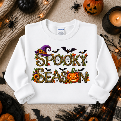 A white long-sleeve crewneck sweatshirt with a 'Spooky Season' graphic, laid flat on a neutral background with Halloween props including pumpkins, a candle, and spider decorations. The brand Gildan is visible on the inner neck label.