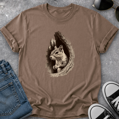 A brown short-sleeve crewneck t-shirt featuring a detailed graphic of a squirrel peeking out of a tree hole on the chest. The shirt is styled in a flat-lay arrangement on a gray concrete-like surface, surrounded by blue jeans, black and white sneakers, and sunglasses.