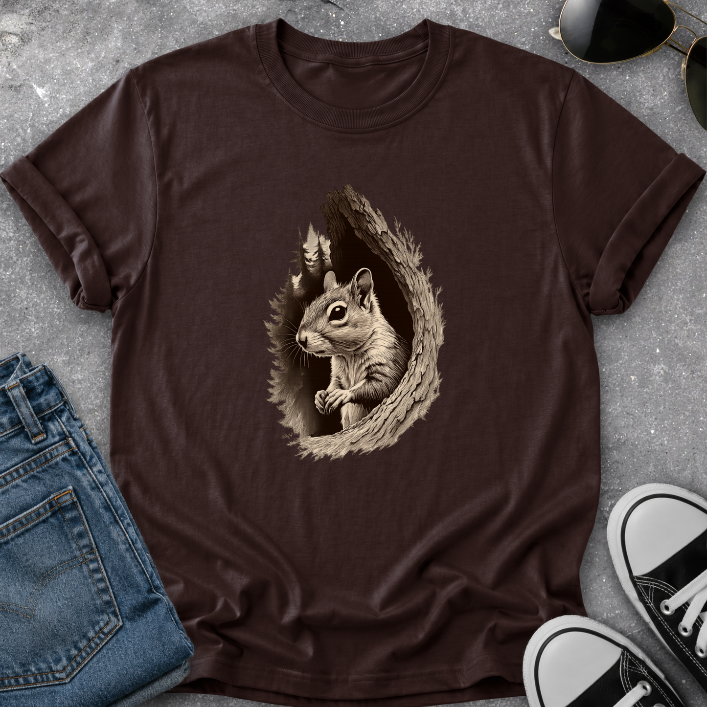 A dark brown short-sleeve t-shirt laid flat on a gray textured surface, featuring a detailed graphic of a squirrel inside a tree hollow. The shirt is styled with blue jeans, sunglasses, and black-and-white sneakers.