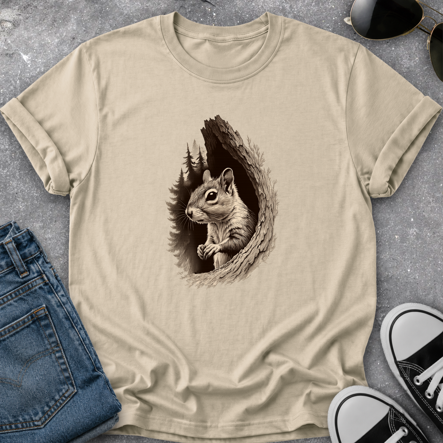 A beige crewneck t-shirt featuring a detailed graphic of a squirrel peeking out of a tree hole, styled in a flat-lay arrangement with blue jeans, sunglasses, and black-and-white sneakers on a gray concrete surface.