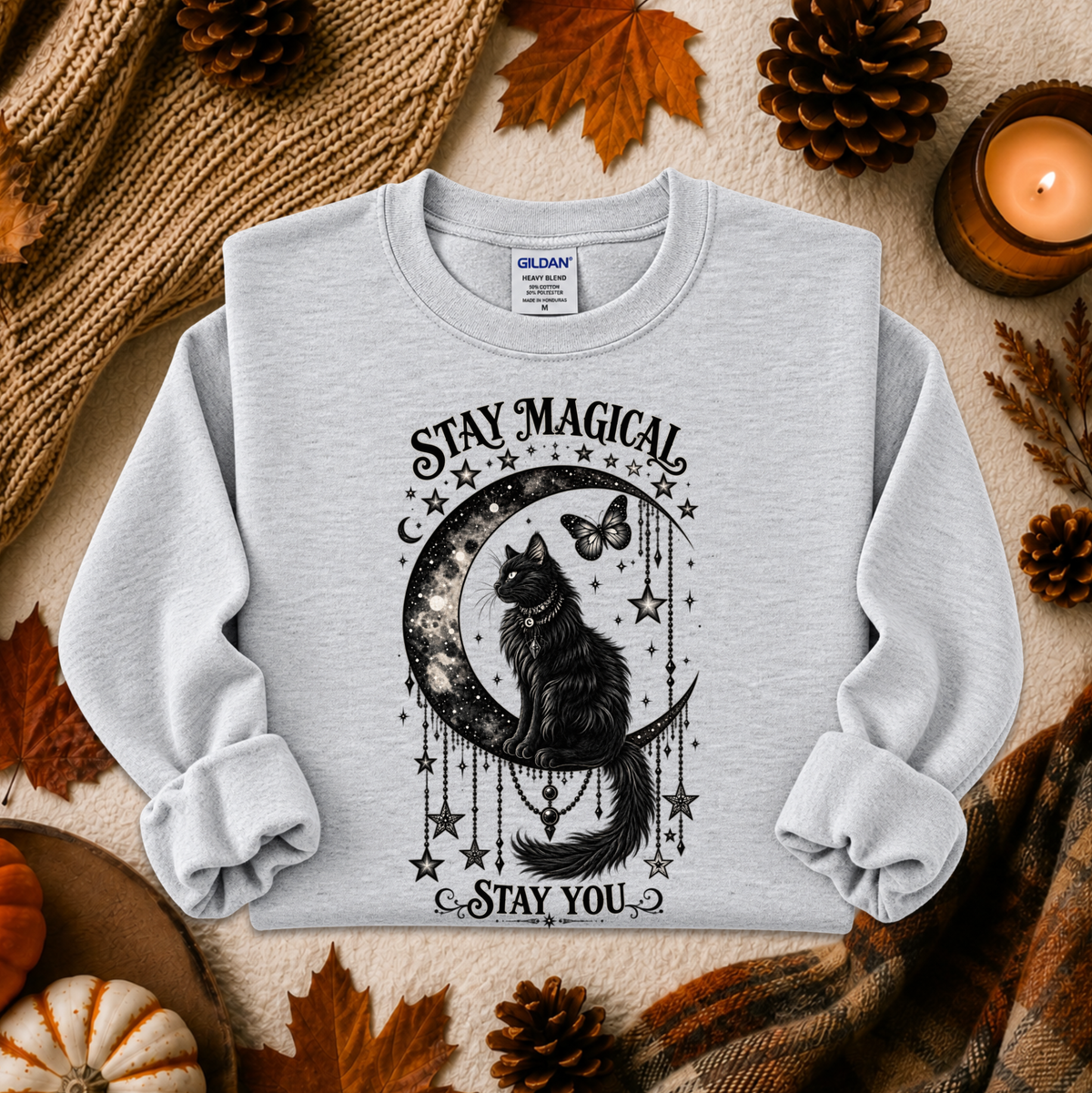 A light heather gray crewneck sweatshirt with a black graphic of a cat on a crescent moon surrounded by stars and the text 'STAY MAGICAL' and 'STAY YOU'. The sweatshirt is displayed in a flat-lay arrangement with autumn props including pinecones, leaves, a candle, and a knit blanket.