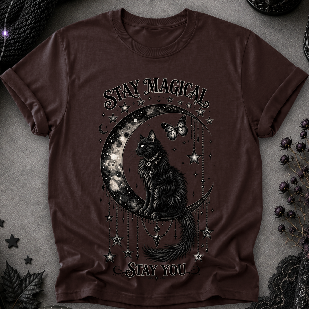 A dark brown short-sleeve t-shirt with a gothic graphic of a black cat sitting on a crescent moon surrounded by stars and a butterfly, with the text 'STAY MAGICAL' and 'STAY YOU'. The shirt is displayed flat on a textured gray surface with dark decorative props.
