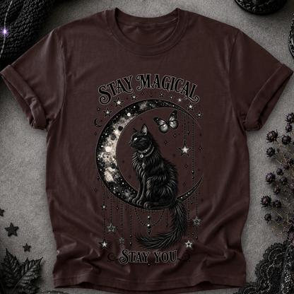 A dark brown short-sleeve t-shirt with a gothic graphic of a black cat sitting on a crescent moon surrounded by stars and a butterfly, with the text 'STAY MAGICAL' and 'STAY YOU'. The shirt is displayed flat on a textured gray surface with dark decorative props.