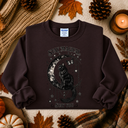 A dark brown crewneck sweatshirt featuring a black graphic of a cat on a crescent moon surrounded by stars, with the text 'Stay Magical' and 'Stay You'. The sweatshirt is styled in a cozy autumn flat-lay with pinecones, a candle, and a knit blanket.