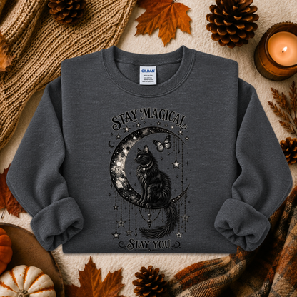 A dark gray crewneck sweatshirt with a black graphic of a cat on a crescent moon surrounded by stars and the text 'STAY MAGICAL' and 'STAY YOU'. The sweatshirt is displayed in a flat-lay autumn setting with pinecones, leaves, a candle, and a knit blanket.