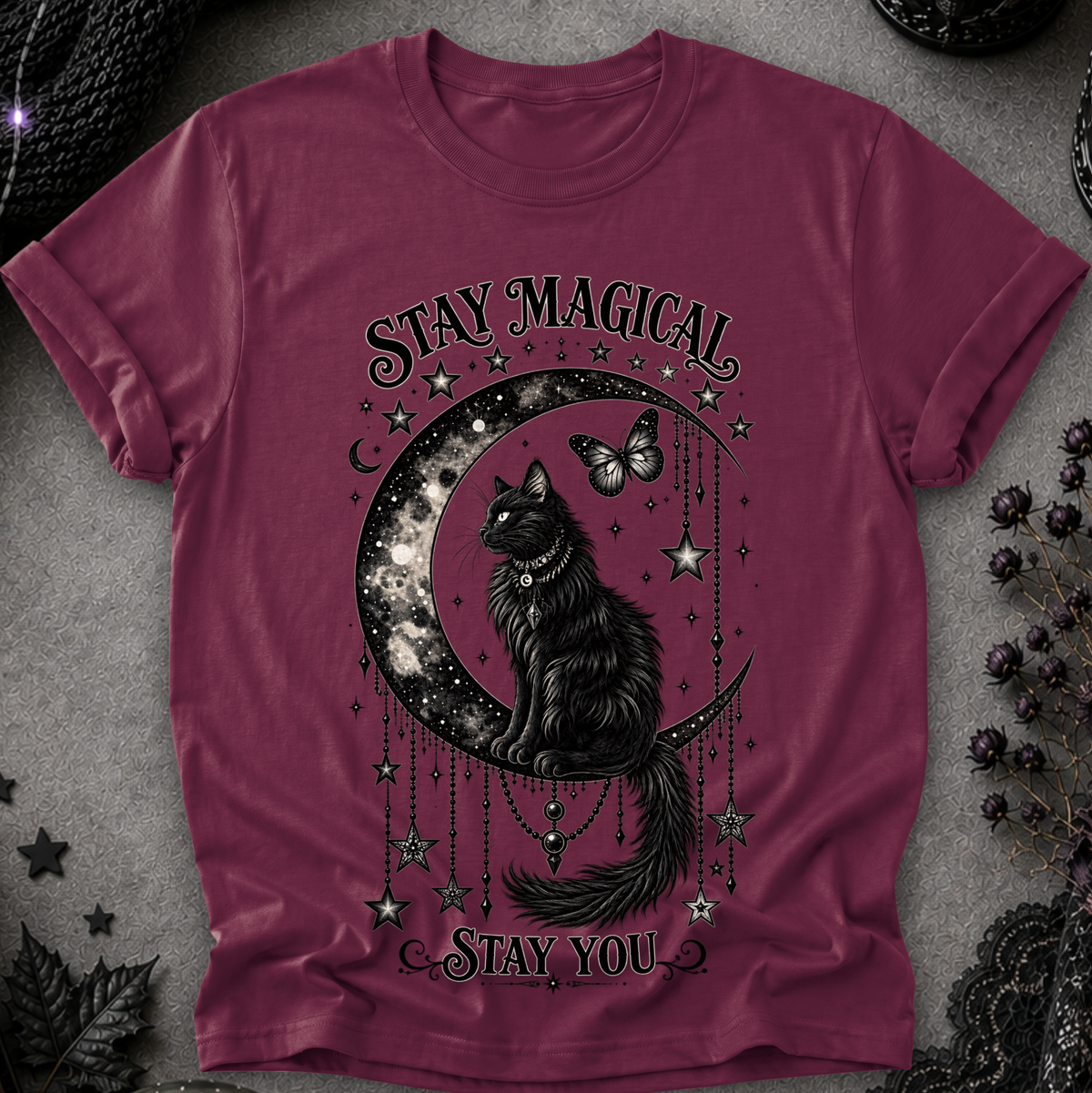 A maroon short-sleeve crewneck t-shirt featuring a gothic graphic of a black cat sitting on a crescent moon surrounded by stars and a butterfly, with the text 'STAY MAGICAL' and 'STAY YOU'. The shirt is laid flat on a textured gray surface with dark decorative props.