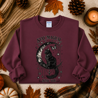 A burgundy crewneck sweatshirt with a black graphic of a cat on a crescent moon surrounded by stars and the words 'STAY MAGICAL' and 'STAY YOU', styled in a flat lay with autumn leaves, pinecones, a candle, and a knit blanket.