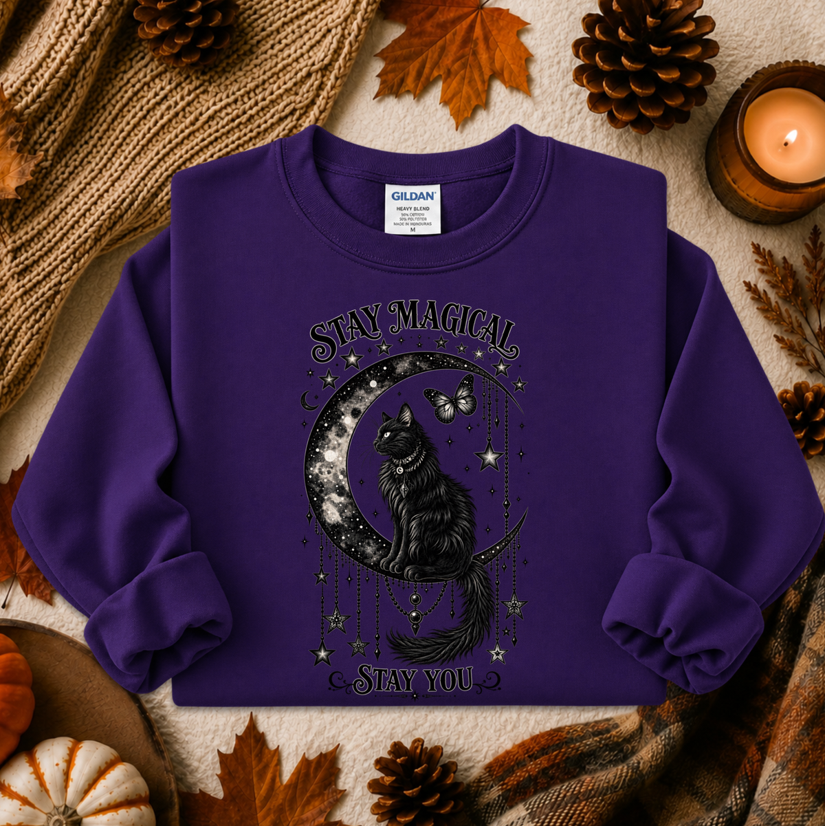 A purple long-sleeve crewneck sweatshirt with a black graphic of a cat on a crescent moon surrounded by stars and the words 'STAY MAGICAL' and 'STAY YOU'. The sweatshirt is displayed in a flat-lay autumn setting with pinecones, leaves, a candle, and a knit blanket.