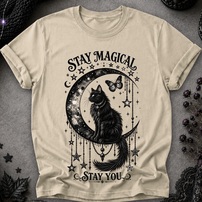 A beige short-sleeve t-shirt with a black graphic print of a cat sitting on a crescent moon surrounded by stars and the text 'STAY MAGICAL' and 'STAY YOU'. The shirt is laid flat on a textured gray surface with dark decorative elements in the corners.