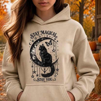 A woman wearing a beige hoodie with a black graphic of a cat on a crescent moon surrounded by stars and the text 'STAY MAGICAL' and 'STAY YOU', photographed outdoors in autumn with pumpkins and fall foliage in the background.