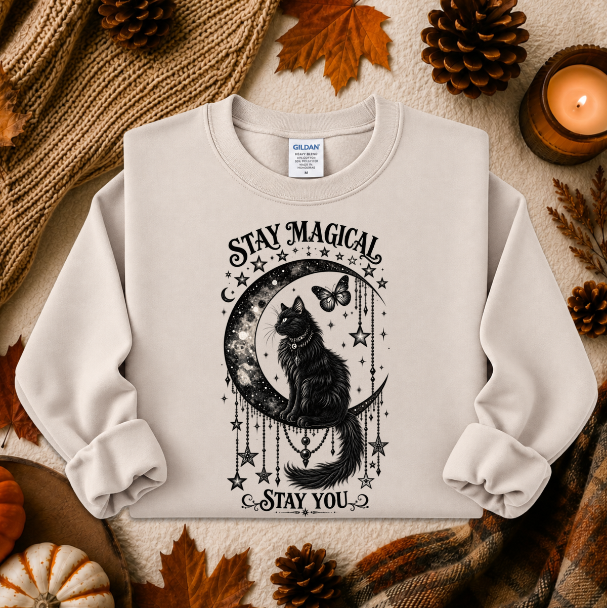 A light beige crewneck sweatshirt with a black graphic of a cat on a crescent moon surrounded by stars and the text 'STAY MAGICAL' and 'STAY YOU'. The sweatshirt is styled in a cozy autumn flat-lay with a knitted blanket, pinecones, leaves, and a lit candle.