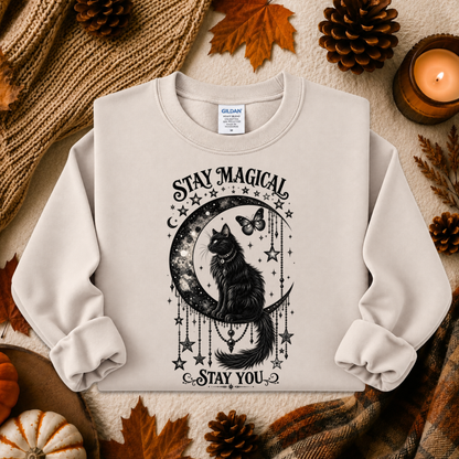 A light beige crewneck sweatshirt with a black graphic of a cat on a crescent moon surrounded by stars and the text 'STAY MAGICAL' and 'STAY YOU'. The sweatshirt is styled in a cozy autumn flat-lay with a knitted blanket, pinecones, leaves, and a lit candle.