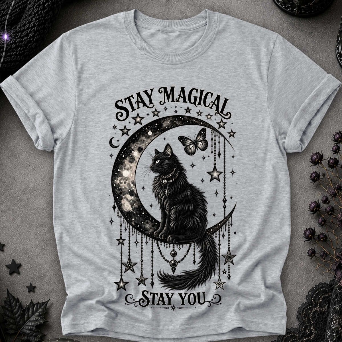 A light heather gray short-sleeve t-shirt with a black graphic print of a cat sitting on a crescent moon surrounded by stars and the text 'STAY MAGICAL' and 'STAY YOU'. The shirt is laid flat on a textured gray surface with dark decorative props in the corners.