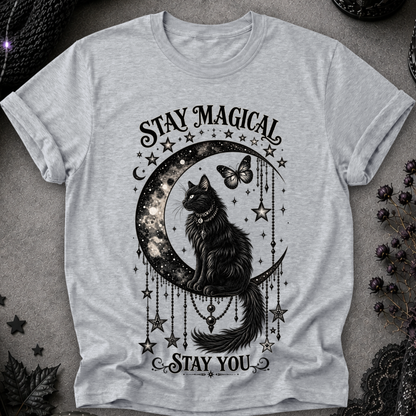 A light heather gray short-sleeve t-shirt with a black graphic print of a cat sitting on a crescent moon surrounded by stars and the text 'STAY MAGICAL' and 'STAY YOU'. The shirt is laid flat on a textured gray surface with dark decorative props in the corners.