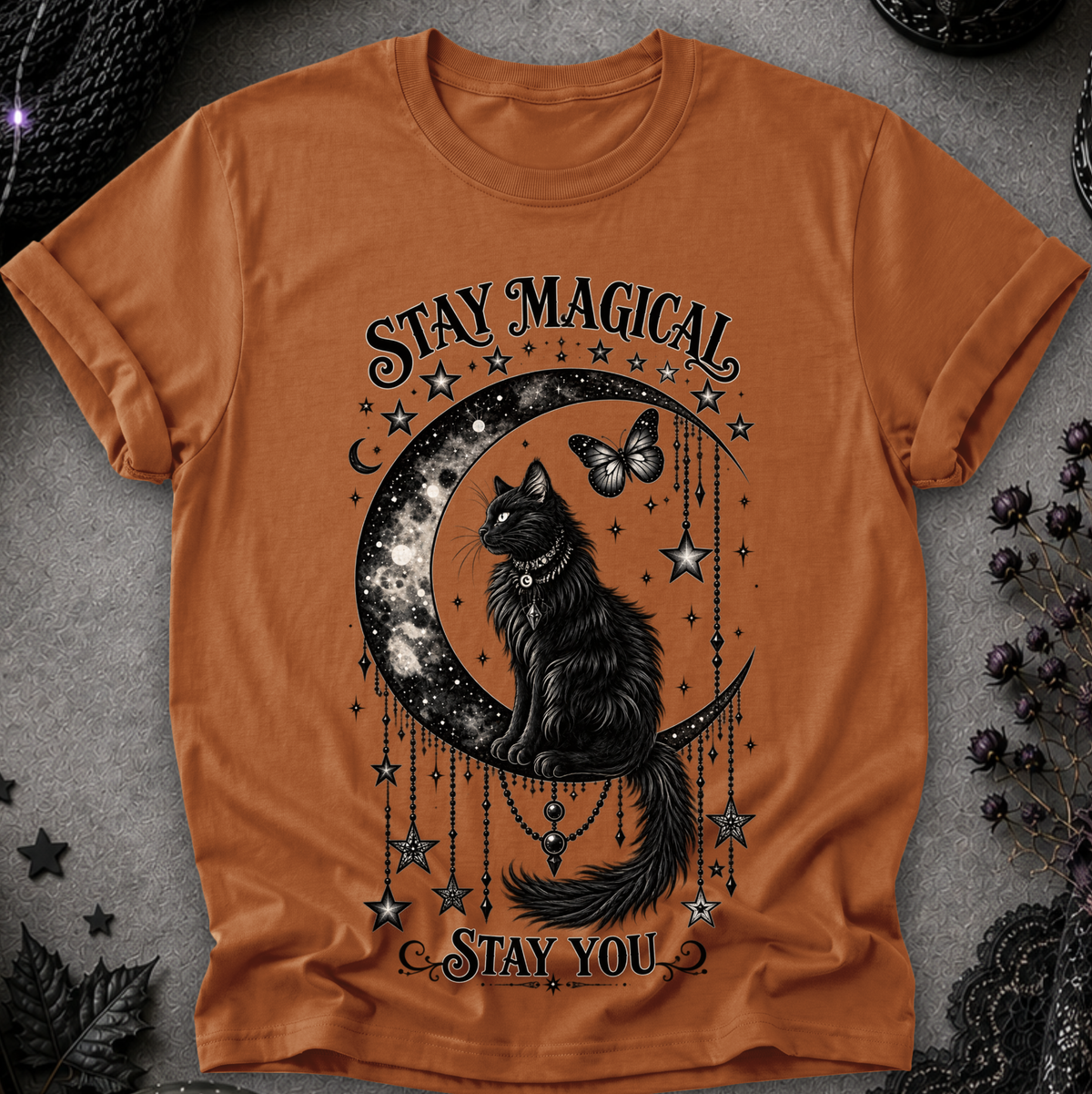 An orange short-sleeve crewneck t-shirt with a gothic graphic of a black cat sitting on a crescent moon surrounded by stars and the text 'STAY MAGICAL' and 'STAY YOU'. The shirt is displayed flat on a textured gray background with dark decorative props.