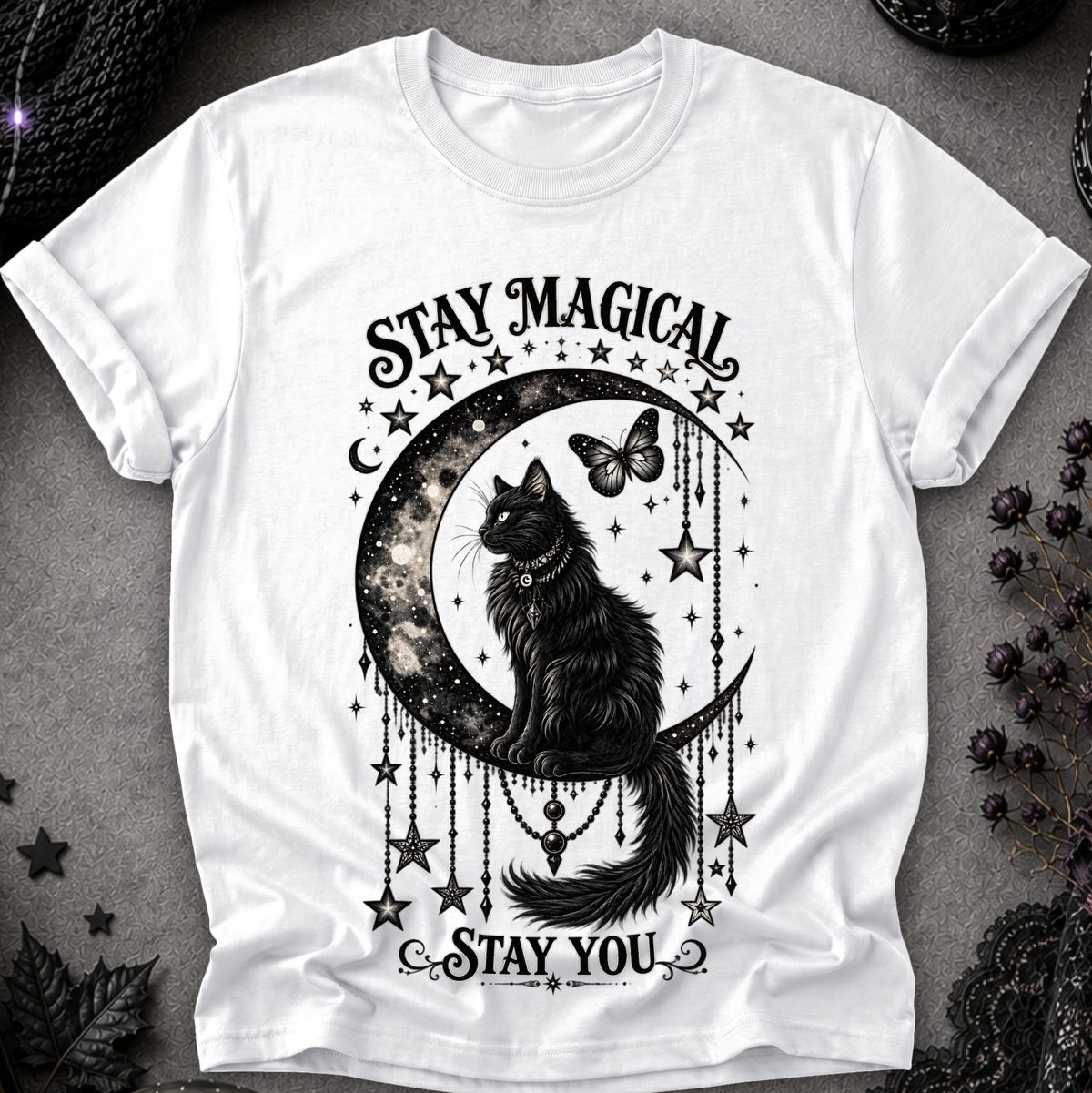 A white short-sleeve crewneck t-shirt featuring a black graphic of a cat sitting on a crescent moon surrounded by stars, with the text 'STAY MAGICAL' above and 'STAY YOU' below, laid flat on a dark textured surface.