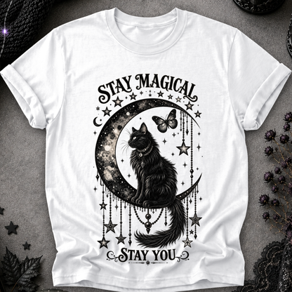 A white short-sleeve crewneck t-shirt featuring a black graphic of a cat sitting on a crescent moon surrounded by stars, with the text 'STAY MAGICAL' above and 'STAY YOU' below, laid flat on a dark textured surface.