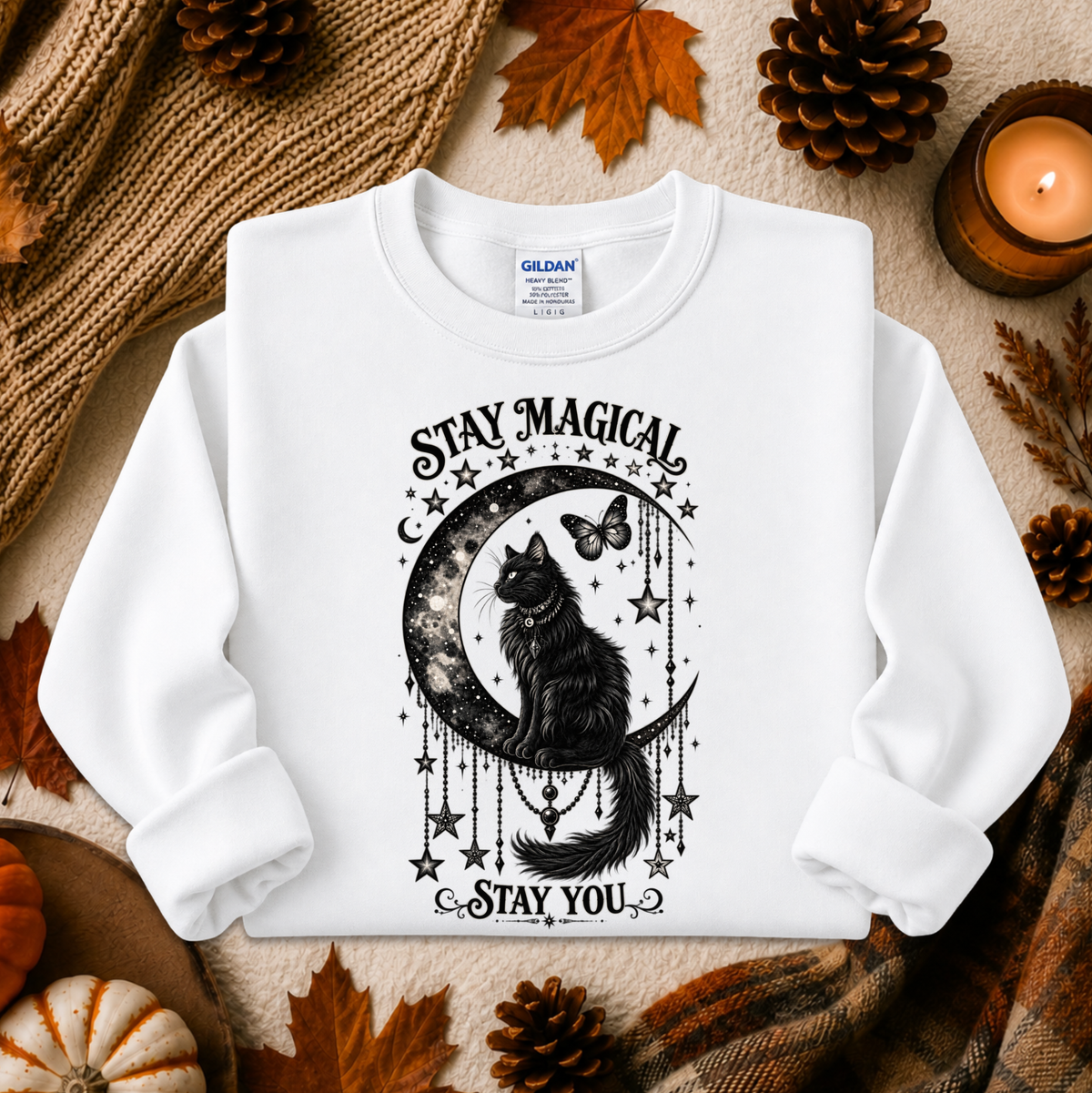 A white long-sleeve crewneck sweatshirt with a black graphic of a cat on a crescent moon surrounded by stars and the text 'STAY MAGICAL' and 'STAY YOU'. The sweatshirt is displayed in a flat-lay arrangement with autumn props including pinecones, a candle, and a knit blanket.