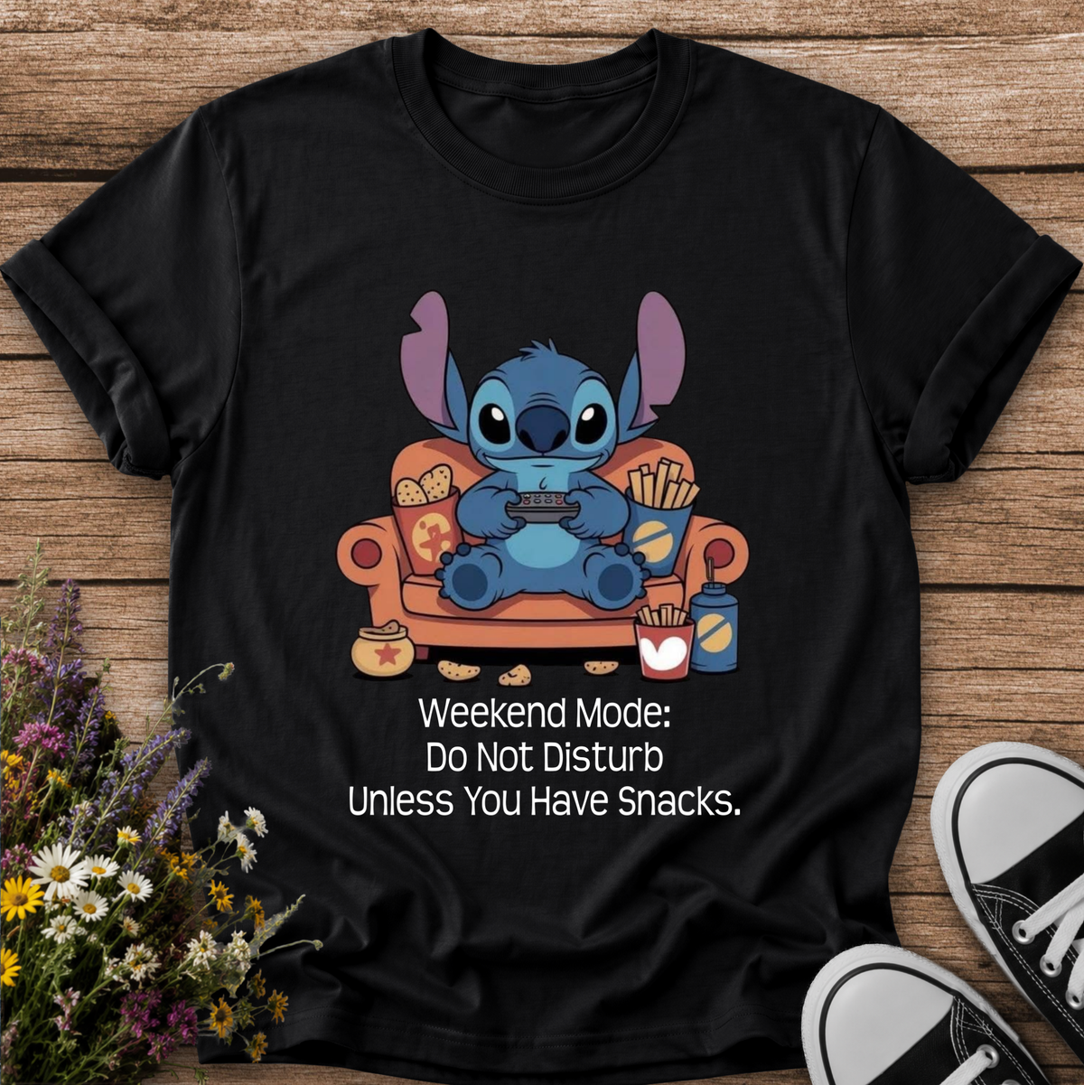 A black short-sleeve graphic T-shirt laid flat on a wooden surface, featuring a cartoon blue character sitting on an orange couch holding a game controller, with the text 'Weekend Mode: Do Not Disturb Unless You Have Snacks.' White flowers and black-and-white sneakers are placed around the shirt as styling props.