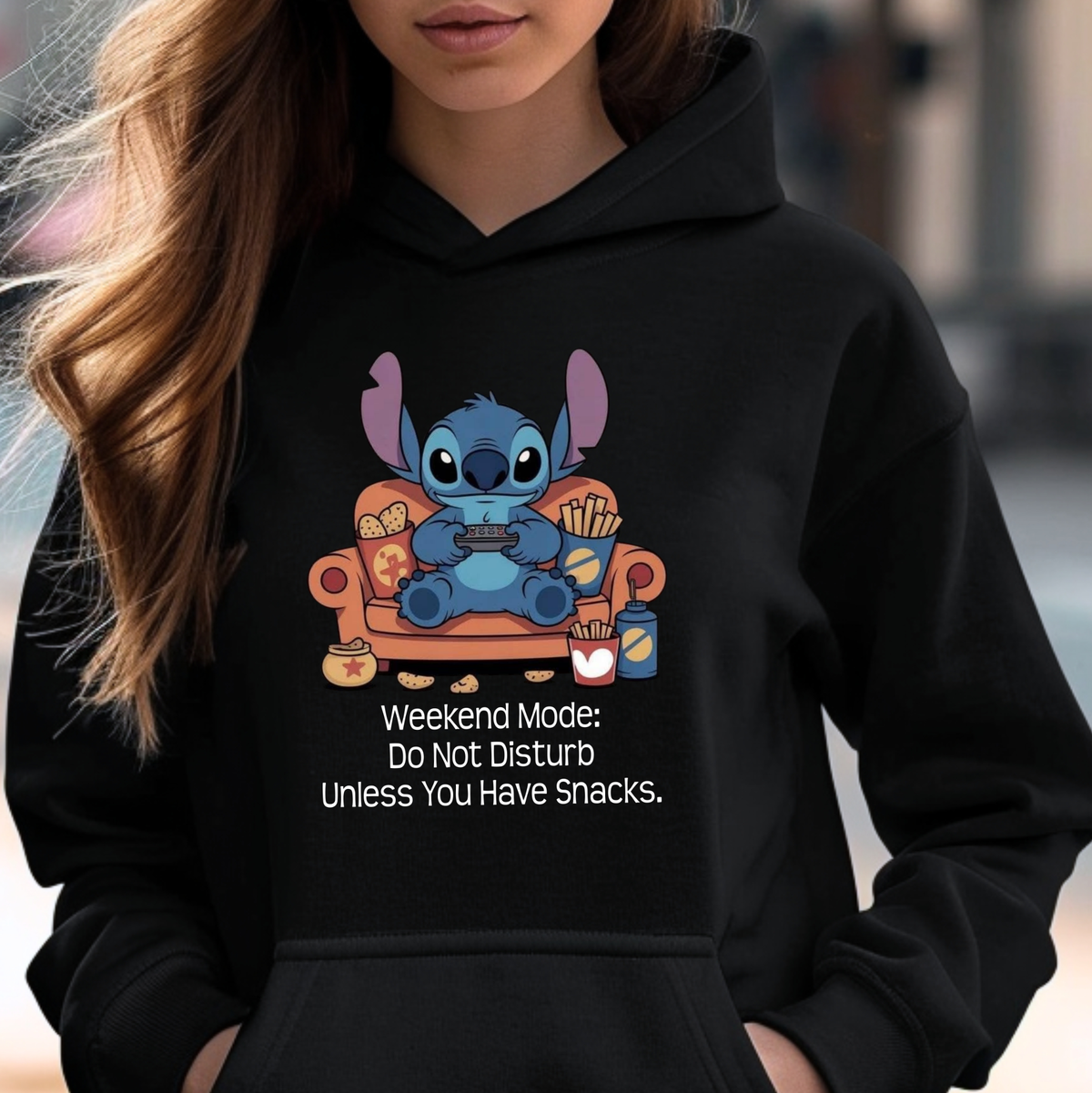 A woman wearing a black hoodie with a cartoon graphic of Stitch sitting on an orange couch holding a game controller, with the text 'Weekend Mode: Do Not Disturb Unless You Have Snacks.' printed below.