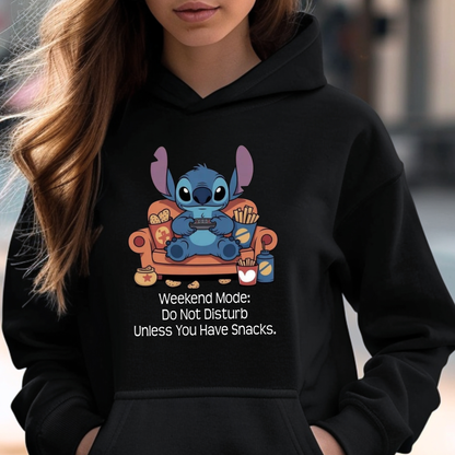 A woman wearing a black hoodie with a cartoon graphic of Stitch sitting on an orange couch holding a game controller, with the text 'Weekend Mode: Do Not Disturb Unless You Have Snacks.' printed below.