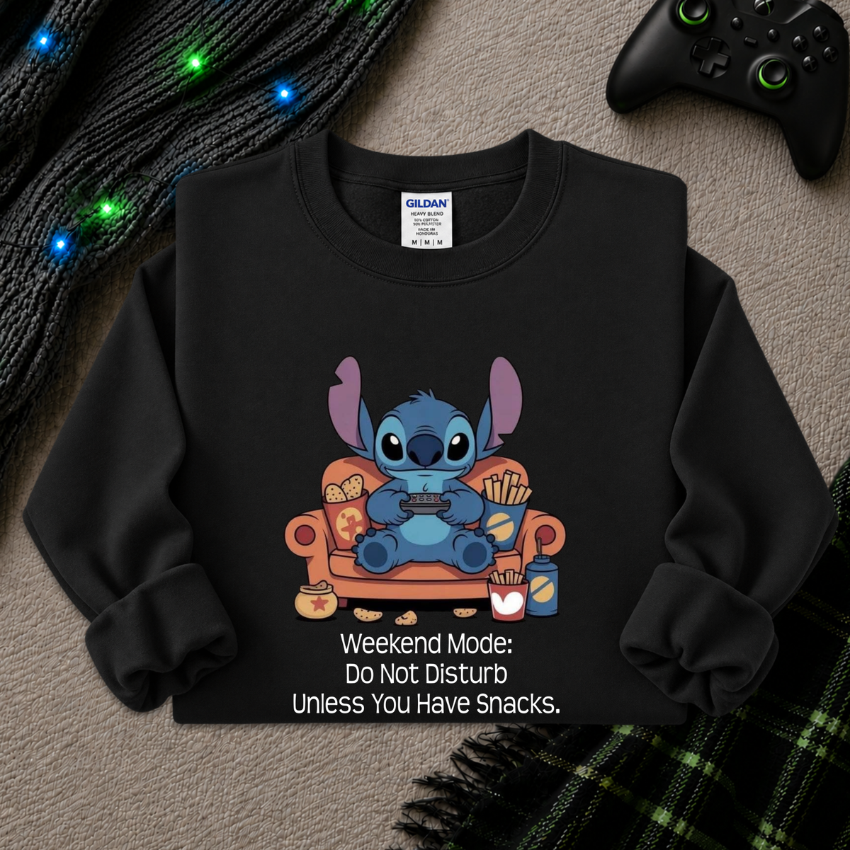 A black long-sleeve sweatshirt laid flat on a textured beige surface, featuring a cartoon character sitting in an orange armchair holding a game controller, with snacks around it and the text 'Weekend Mode: Do Not Disturb Unless You Have Snacks.' A Gildan neck label is visible, and the sweatshirt is styled with a black knit blanket, glowing blue lights, and a black game controller.