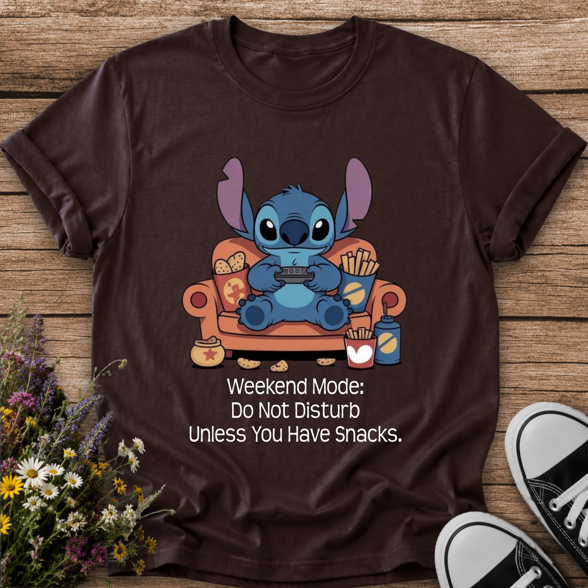 A dark brown graphic t-shirt featuring the character Stitch sitting on an orange couch with snacks, with the text 'Weekend Mode: Do Not Disturb Unless You Have Snacks.' printed below.