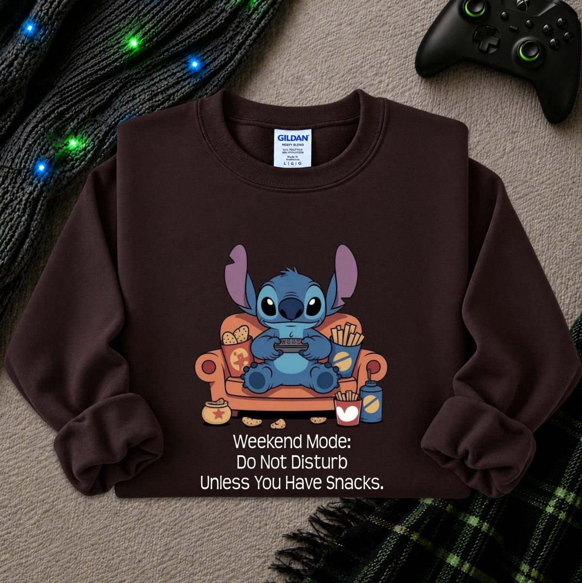 A dark brown crewneck sweatshirt featuring a cartoon graphic of Stitch sitting on an orange couch eating snacks, with the text 'Weekend Mode: Do Not Disturb Unless You Have Snacks.' printed below.