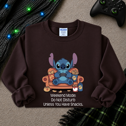 A dark brown crewneck sweatshirt featuring a cartoon graphic of Stitch sitting on an orange couch eating snacks, with the text 'Weekend Mode: Do Not Disturb Unless You Have Snacks.' printed below.