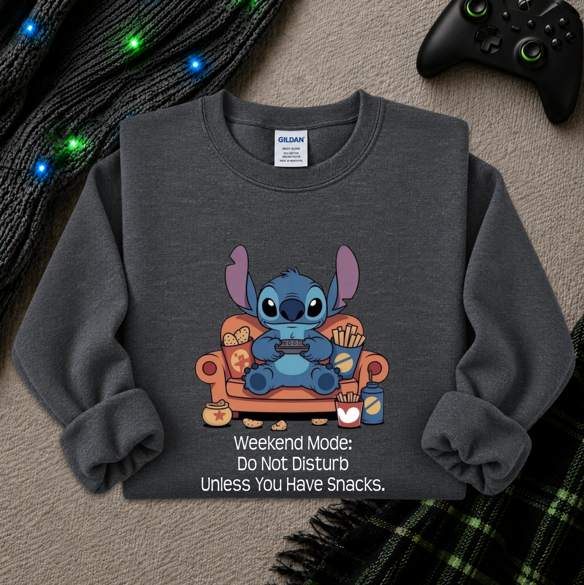 A dark gray crewneck sweatshirt featuring a cartoon graphic of Stitch sitting on an orange couch holding a game controller, with the text 'Weekend Mode: Do Not Disturb Unless You Have Snacks.' printed below.