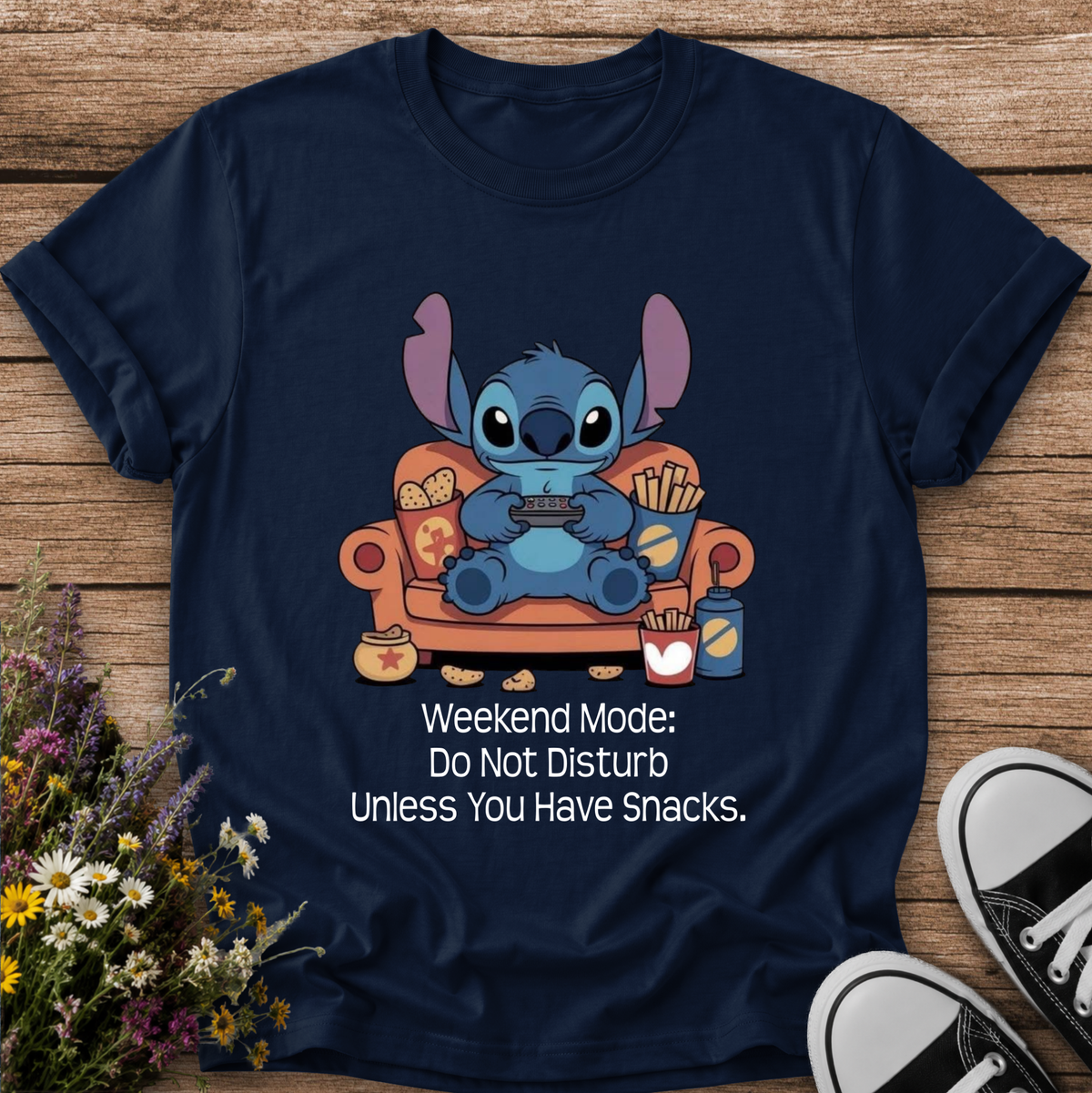 A navy blue graphic t-shirt featuring a cartoon illustration of Stitch sitting on an orange couch eating snacks, with the text 'Weekend Mode: Do Not Disturb Unless You Have Snacks.' printed below.