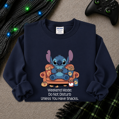 A navy blue crewneck sweatshirt featuring a cartoon graphic of Stitch sitting on an orange couch holding a game controller, with the text 'Weekend Mode: Do Not Disturb Unless You Have Snacks.' printed below.