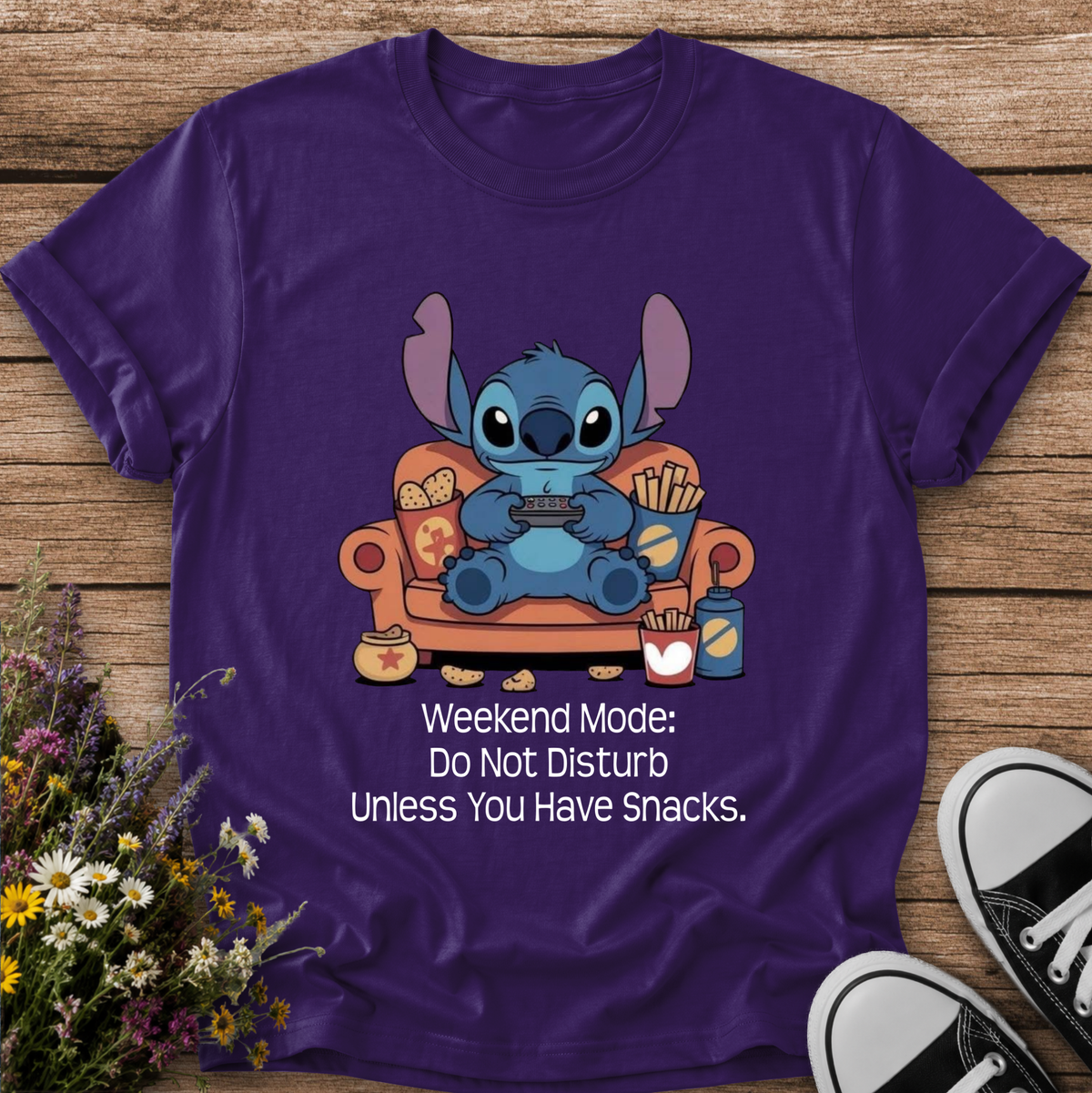 A purple short-sleeve graphic T-shirt laid flat on a wooden surface, featuring a cartoon blue character sitting on an orange couch with snacks and the text 'Weekend Mode: Do Not Disturb Unless You Have Snacks.' Wildflowers and black-and-white sneakers are placed around the shirt as styling props.