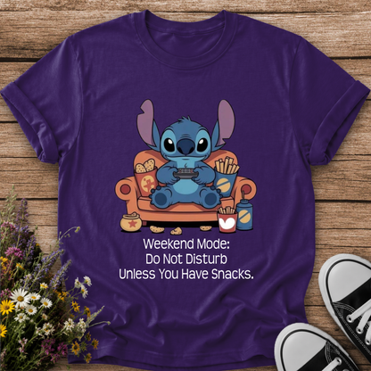 A purple short-sleeve graphic T-shirt laid flat on a wooden surface, featuring a cartoon blue character sitting on an orange couch with snacks and the text 'Weekend Mode: Do Not Disturb Unless You Have Snacks.' Wildflowers and black-and-white sneakers are placed around the shirt as styling props.