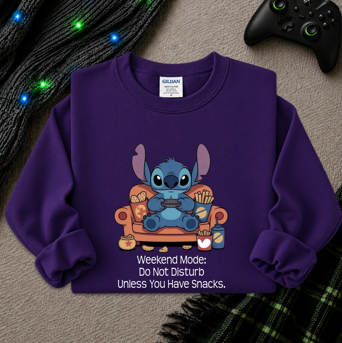 A purple long-sleeve sweatshirt laid flat on a textured gray surface, featuring a cartoon character sitting on an orange couch with snacks and the text 'Weekend Mode: Do Not Disturb Unless You Have Snacks.' A Gildan brand tag is visible inside the collar.