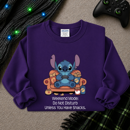 A purple long-sleeve sweatshirt laid flat on a textured gray surface, featuring a cartoon character sitting on an orange couch with snacks and the text 'Weekend Mode: Do Not Disturb Unless You Have Snacks.' A Gildan brand tag is visible inside the collar.