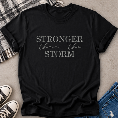 A black short-sleeve graphic T-shirt laid flat on a light gray surface, styled with a plaid blanket, black-and-white sneakers, and blue jeans. The shirt has the phrase “STRONGER than the STORM” printed in white across the chest.