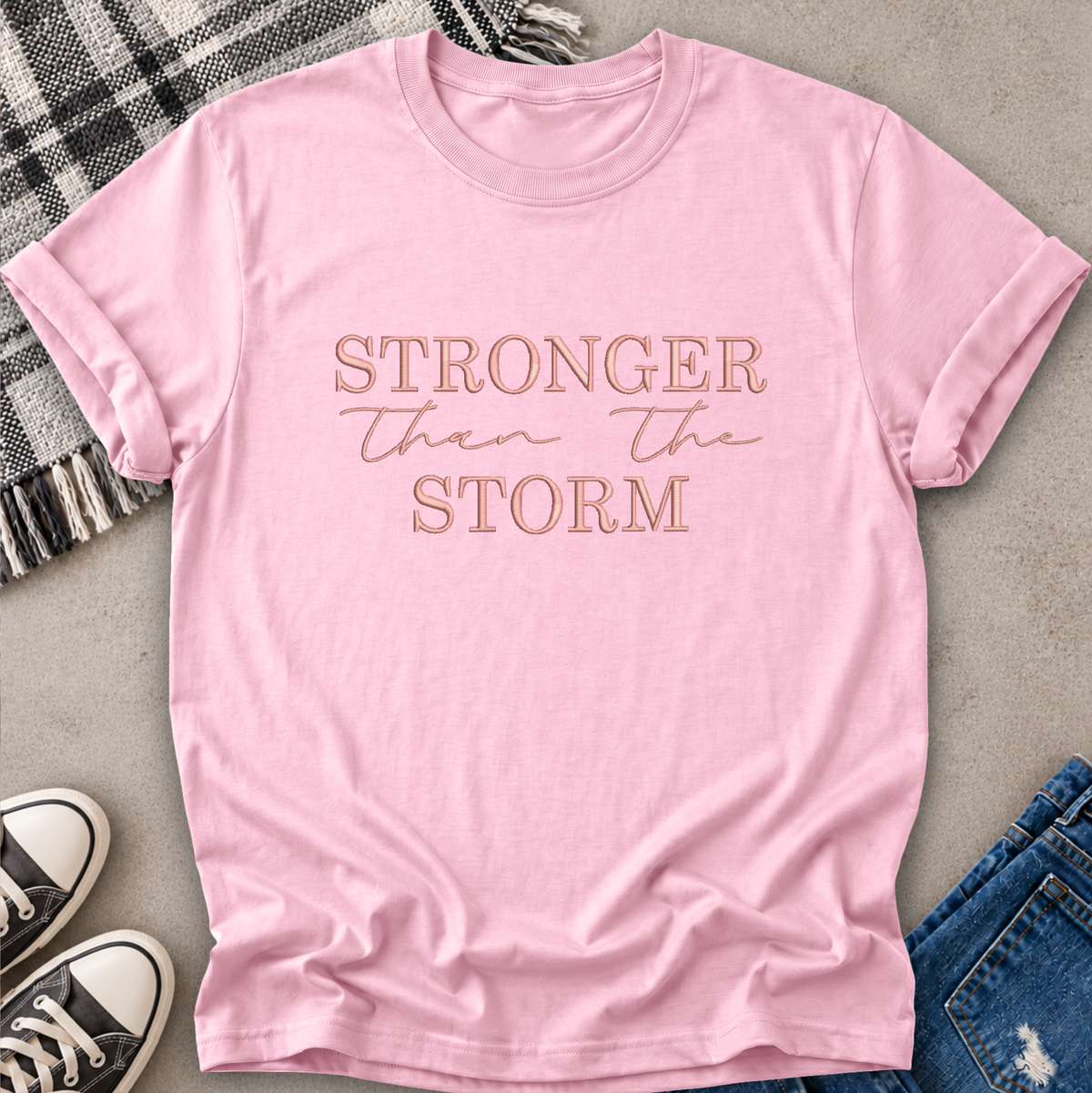 A light pink short-sleeve t-shirt with the phrase 'STRONGER than the STORM' printed in rose gold lettering across the chest, styled in a flat lay with jeans, sneakers, and a plaid blanket on a gray concrete background.