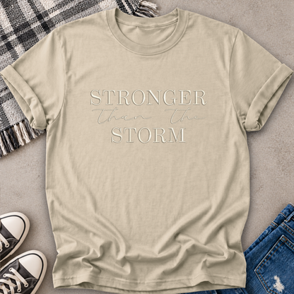 A beige graphic t-shirt laid flat on a gray textured surface, styled with a plaid blanket, black sneakers, and blue jeans. The t-shirt features the text 'STRONGER than the STORM' printed in a light beige/white tone across the chest.