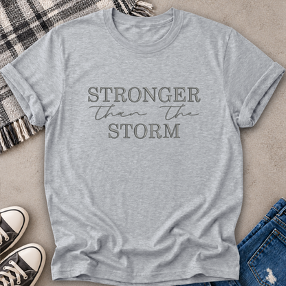 A light heather gray short-sleeve t-shirt with the text 'STRONGER than the STORM' printed on the front in a dark gray tone. The shirt is styled in a flat-lay arrangement on a textured beige surface, surrounded by a black and white plaid blanket, black and white sneakers, and blue jeans.