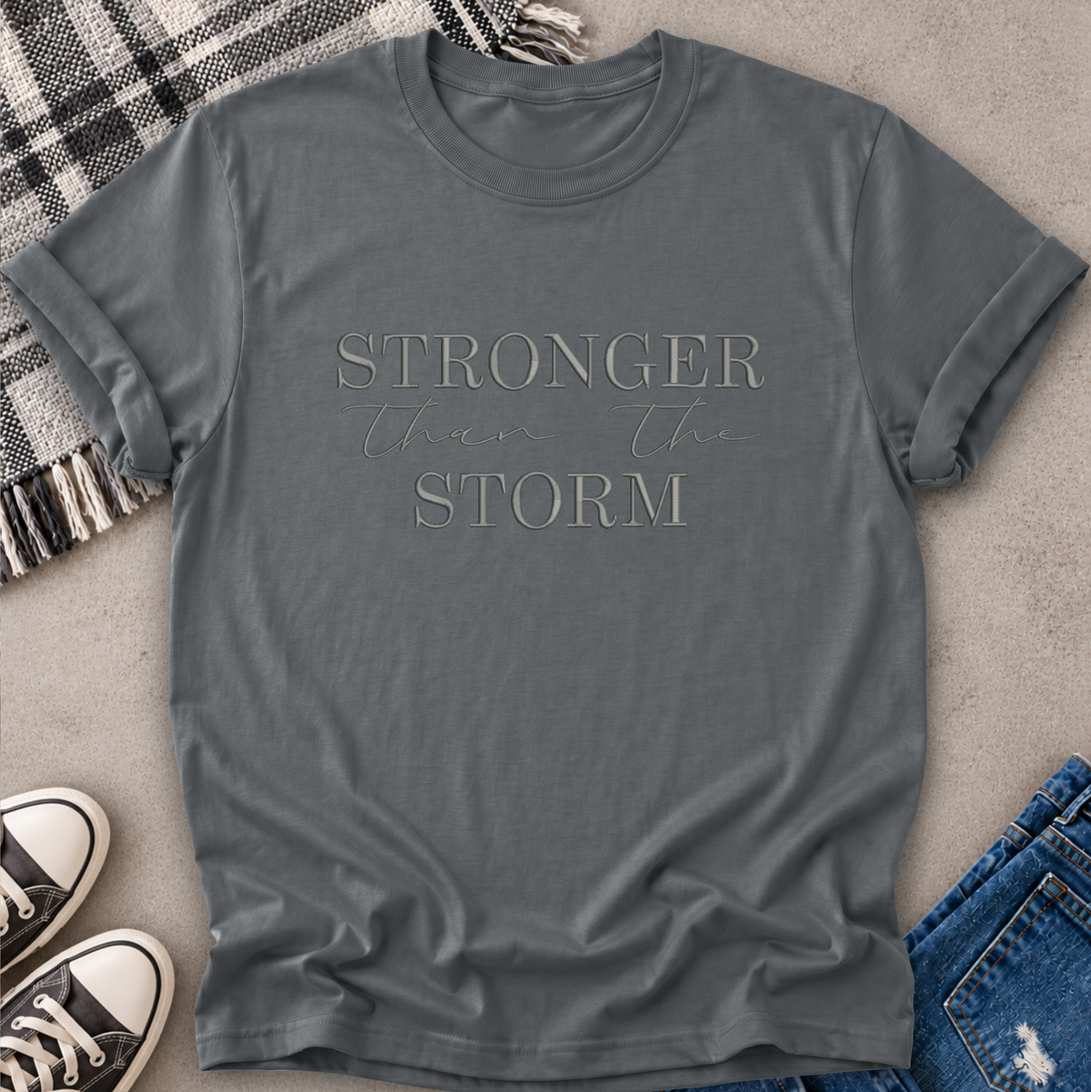A slate gray crewneck t-shirt with the text 'STRONGER than the STORM' printed on the chest, styled in a flat lay with jeans, sneakers, a plaid blanket, and a concrete floor background.