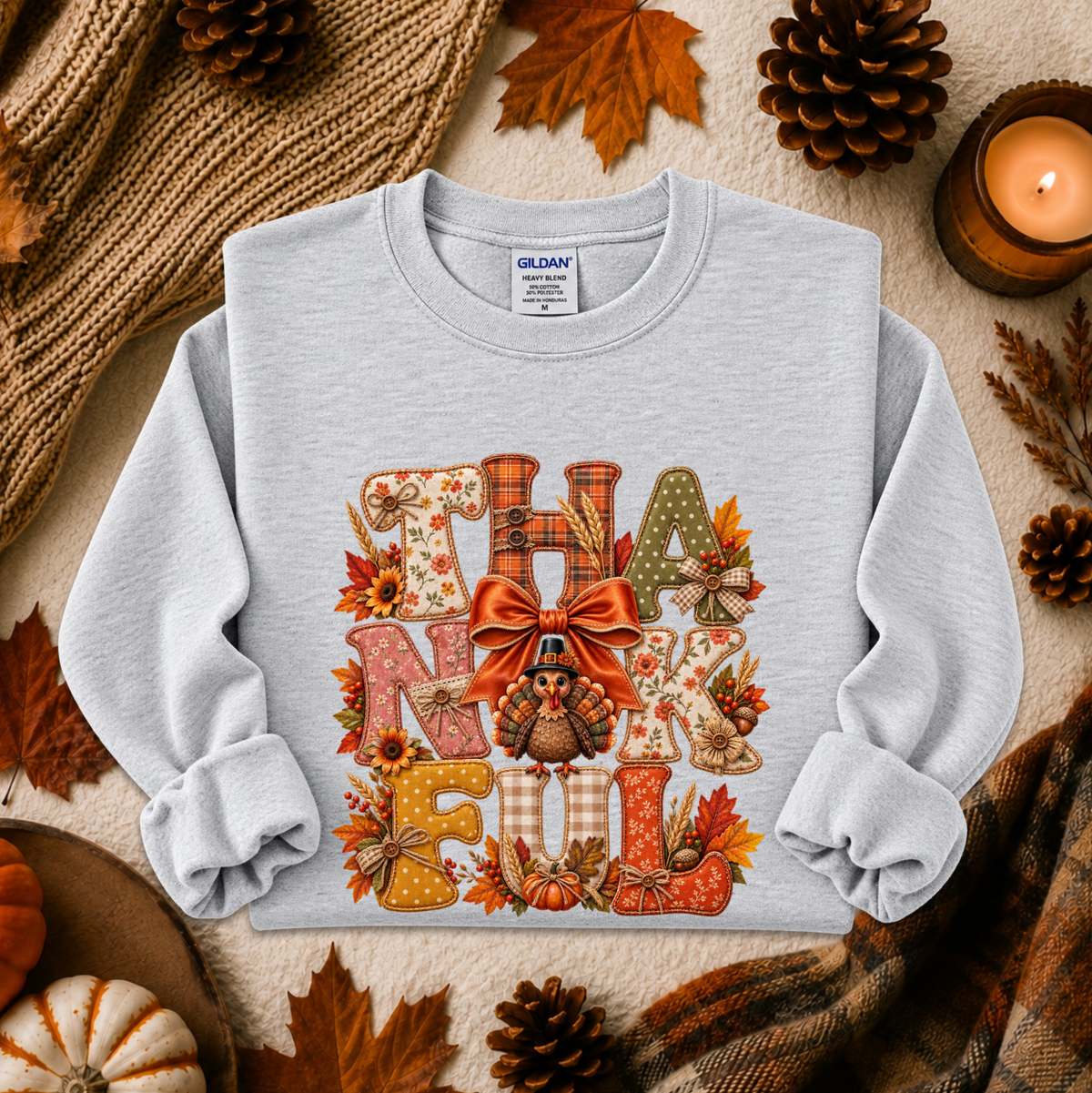 A light heather gray long-sleeve sweatshirt with a large 'THANKsgiving' autumn-themed graphic on the front, styled in a flat lay with a knitted blanket, candles, pinecones, and fall leaves.