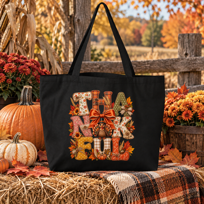 A black canvas tote bag with a large 'THANKFUL' lettering design featuring autumn motifs, displayed on a hay bale with pumpkins, flowers, and a plaid blanket in an outdoor fall setting.