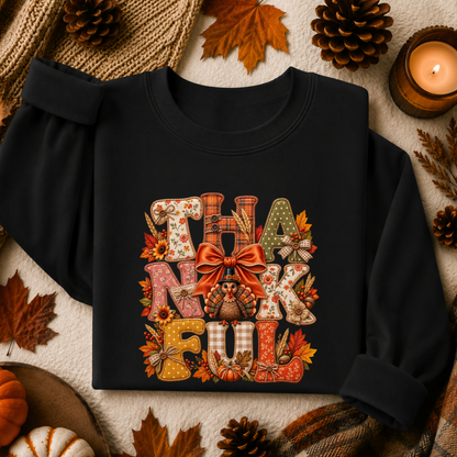 A black long-sleeve sweatshirt laid flat with rolled cuffs, featuring a large autumn-themed 'THANKFUL' graphic on the front. The design includes patterned letters, a red bow, pumpkins, and fall leaves.
