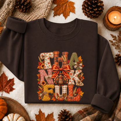 A dark brown crewneck sweatshirt with a festive 'THANKFUL' graphic printed on the front, styled in a cozy autumn flat-lay with pinecones, leaves, and a candle.