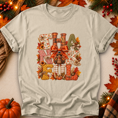 A light beige graphic t-shirt featuring a 'THANKsgiving' design with autumn-themed letters, a turkey, and a bow. The shirt is styled in a flat-lay with seasonal props like a small pumpkin, pine branches, and fall leaves.